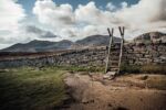 stone fence with wooden ladder propped against it and blue sky, clouds and hills in background.