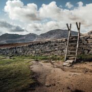 stone fence with wooden ladder propped against it and blue sky, clouds and hills in background.