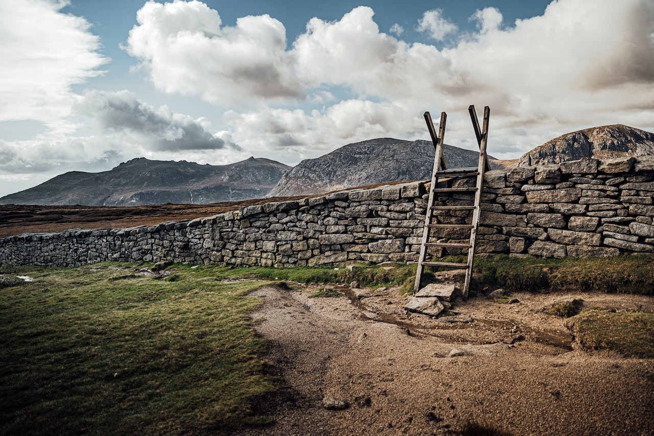stone fence with wooden ladder propped against it and blue sky, clouds and hills in background.