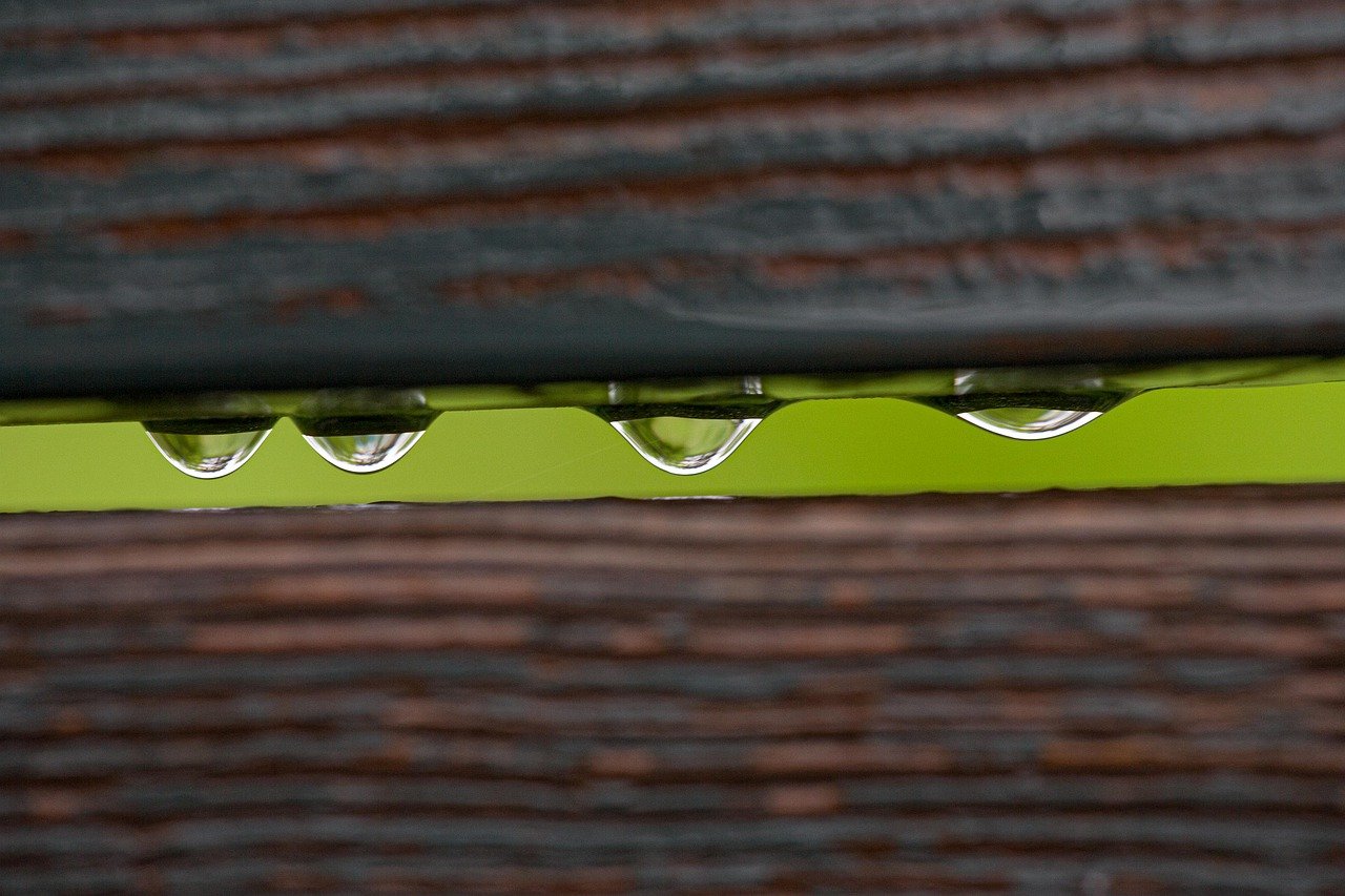 two small horizontal wood fence slabs with a small opening and four drops of water between them and bright green in the background seen through the opening
