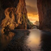 A smooth river surrounded by rock walls leading to an opening through a stone arch at sunset.