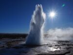 Geyser erupting against a bright blue sky and sun