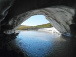 From the inside of a white cave within which runs a body of water, a view of green hills and blue sky