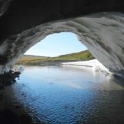 From the inside of a white cave within which runs a body of water, a view of green hills and blue sky