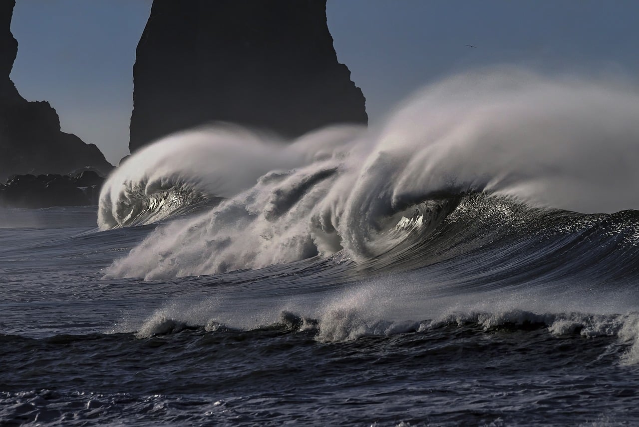 A large wave crashing on the shore with a tall rock in the background.