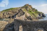A beautiful arched bridge, over the sea, made of rock connecting to a rocky green mountain.