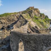 A beautiful arched bridge, over the sea, made of rock connecting to a rocky green mountain.