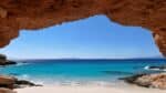 Looking out from a beautiful red rock cave to a beach with blue water and sky.