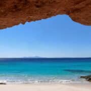 Looking out from a beautiful red rock cave to a beach with blue water and sky.