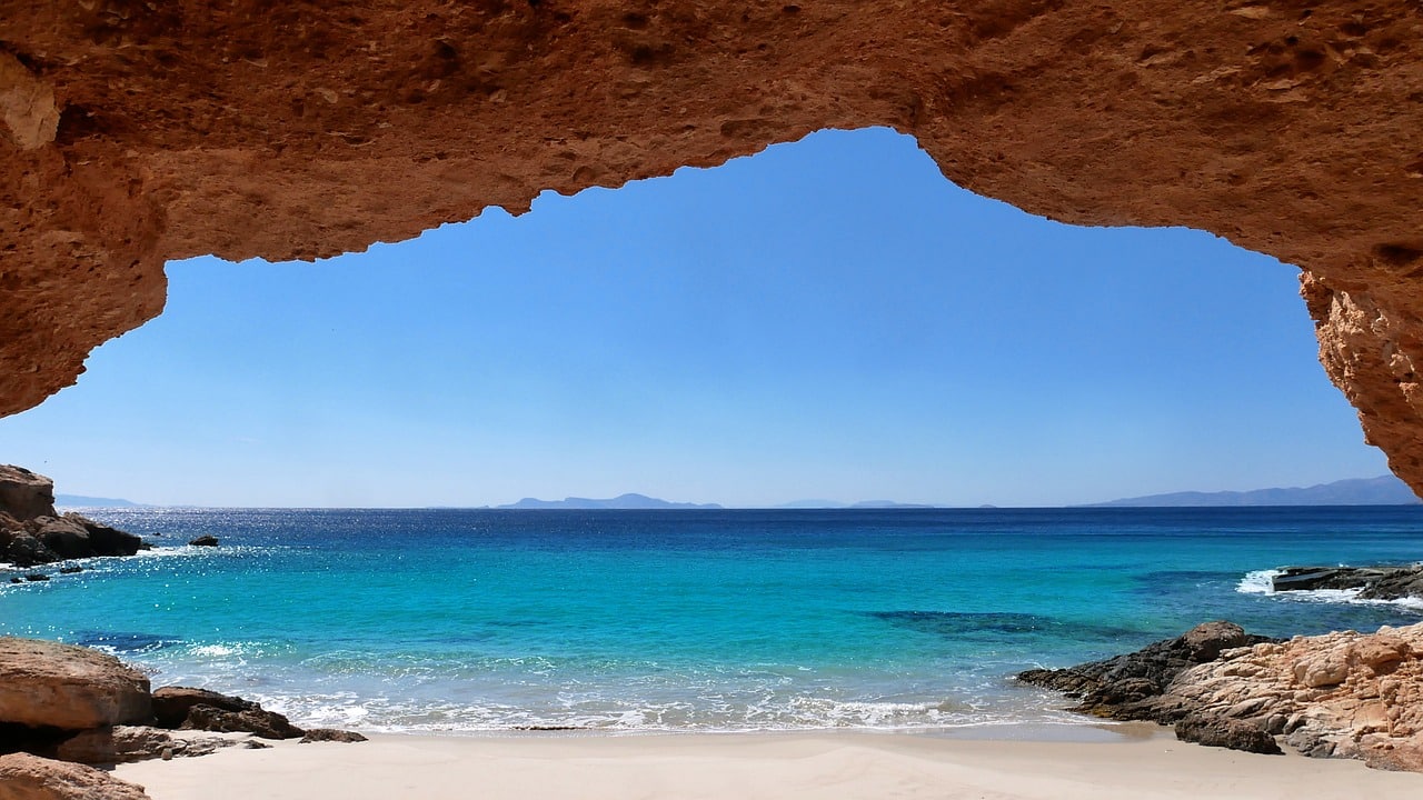 Looking out from a beautiful red rock cave to a beach with blue water and sky.