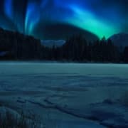 A beautiful scene on a partly frozen lake with mountains and brilliant blue and green northern lights on a dark night.