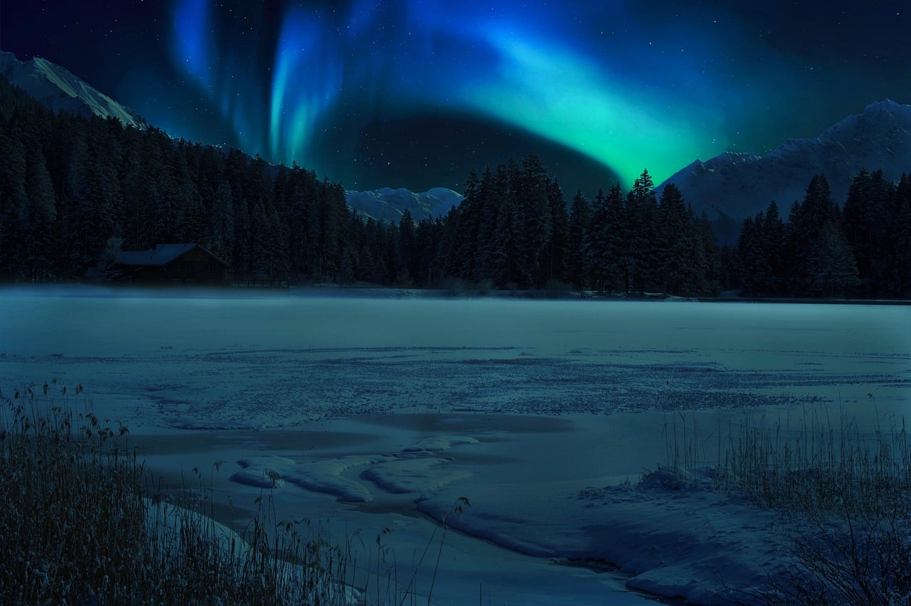 A beautiful scene on a partly frozen lake with mountains and brilliant blue and green northern lights on a dark night.