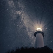 A photo of a lighthouse with a strong light, taken at an angle from the ground with a background of stars in the nighht sky.
