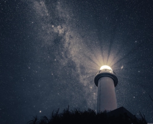 A photo of a lighthouse with a strong light, taken at an angle from the ground with a background of stars in the nighht sky.