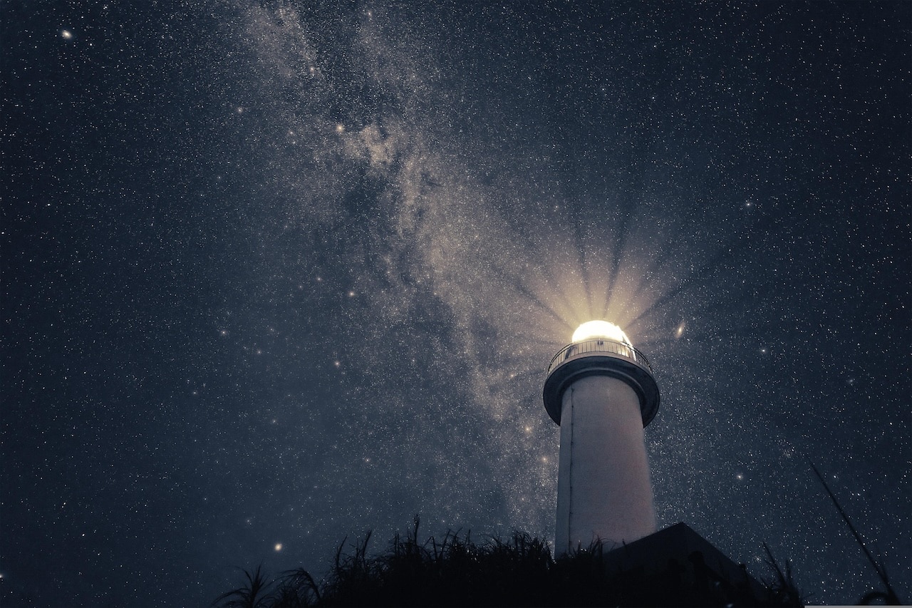 A photo of a lighthouse with a strong light, taken at an angle from the ground with a background of stars in the nighht sky.