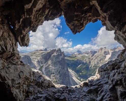 Looking out from inside a cave 's heart shaped opening to mountains, clouds and blue sky.