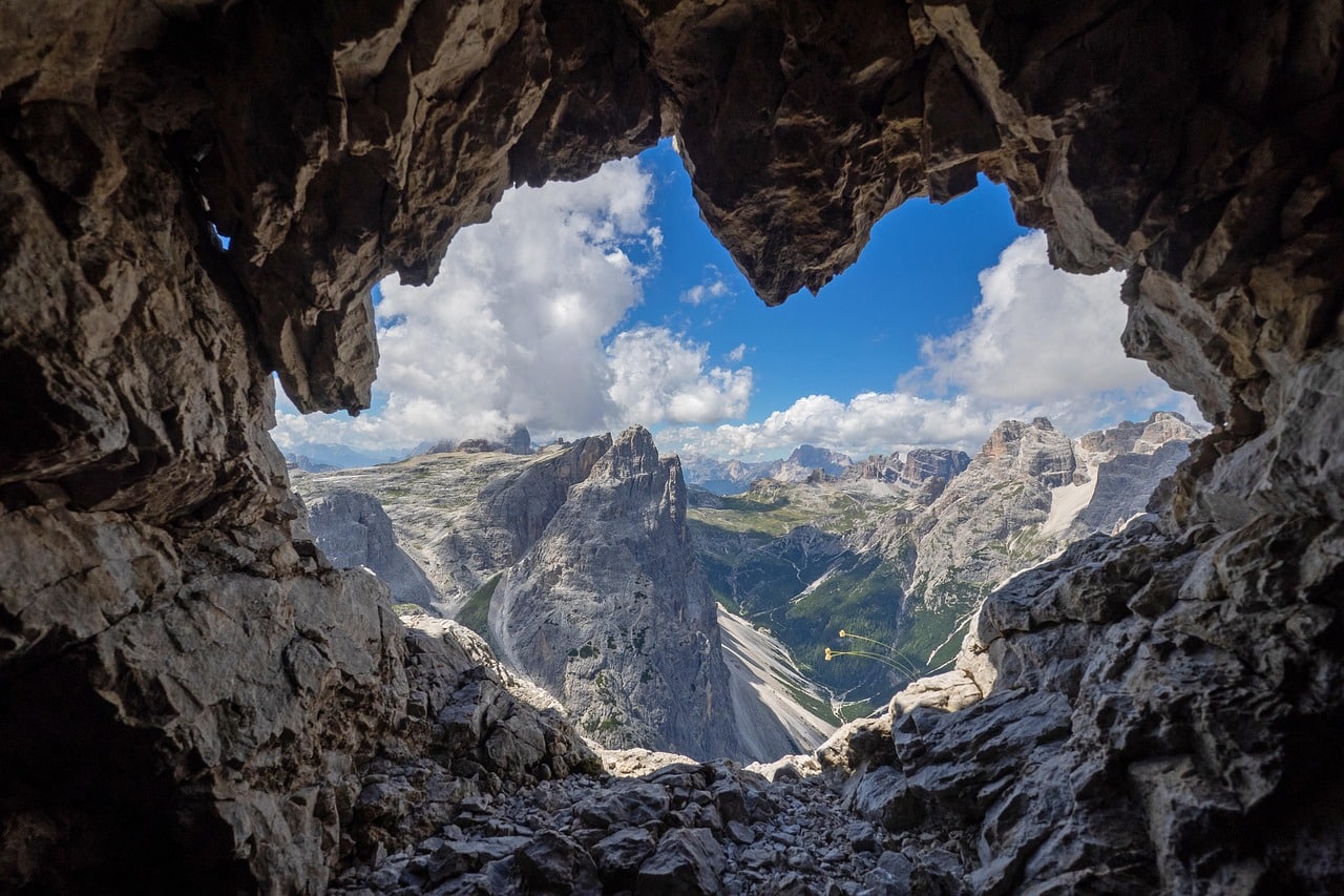 Looking out from inside a cave 's heart shaped opening to mountains, clouds and blue sky.